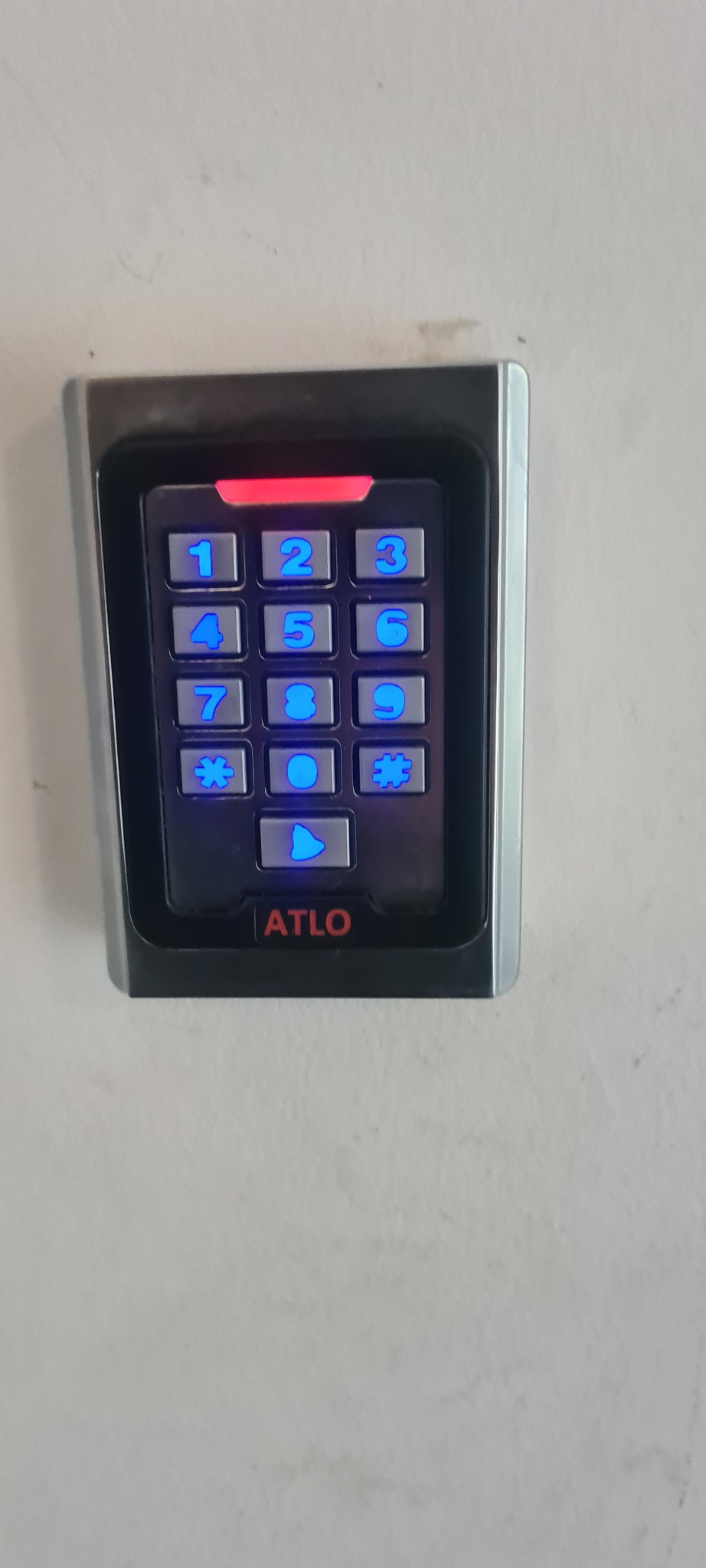 Zamontowany, podświetlany panel domofonu ATLO z klawiaturą numeryczną na jasnej ścianie, widok z bliska.