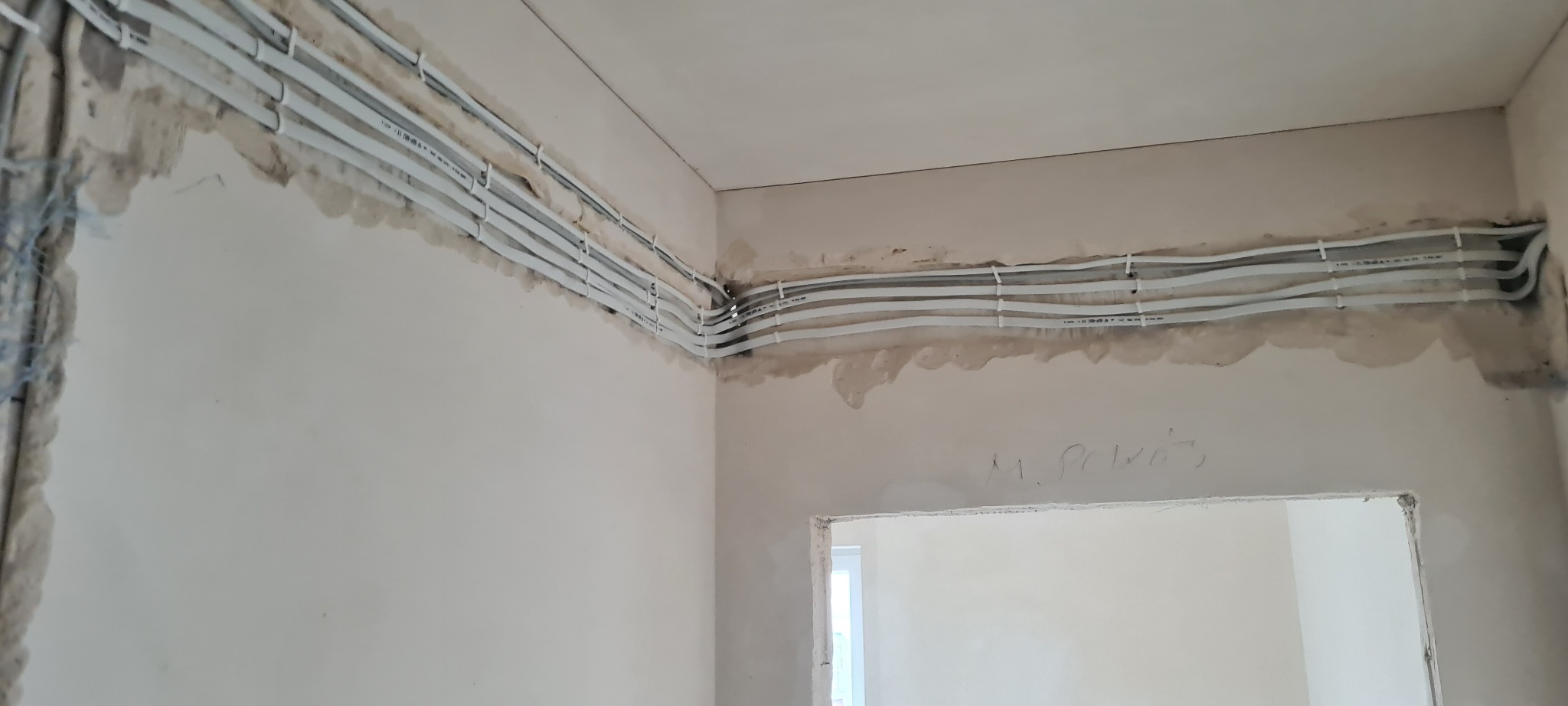 Białe kable elektryczne ułożone równolegle pod sufitem w surowym wnętrzu. Widoczne mocowania kabli do ściany. Ściany w trakcie prac wykończeniowych.