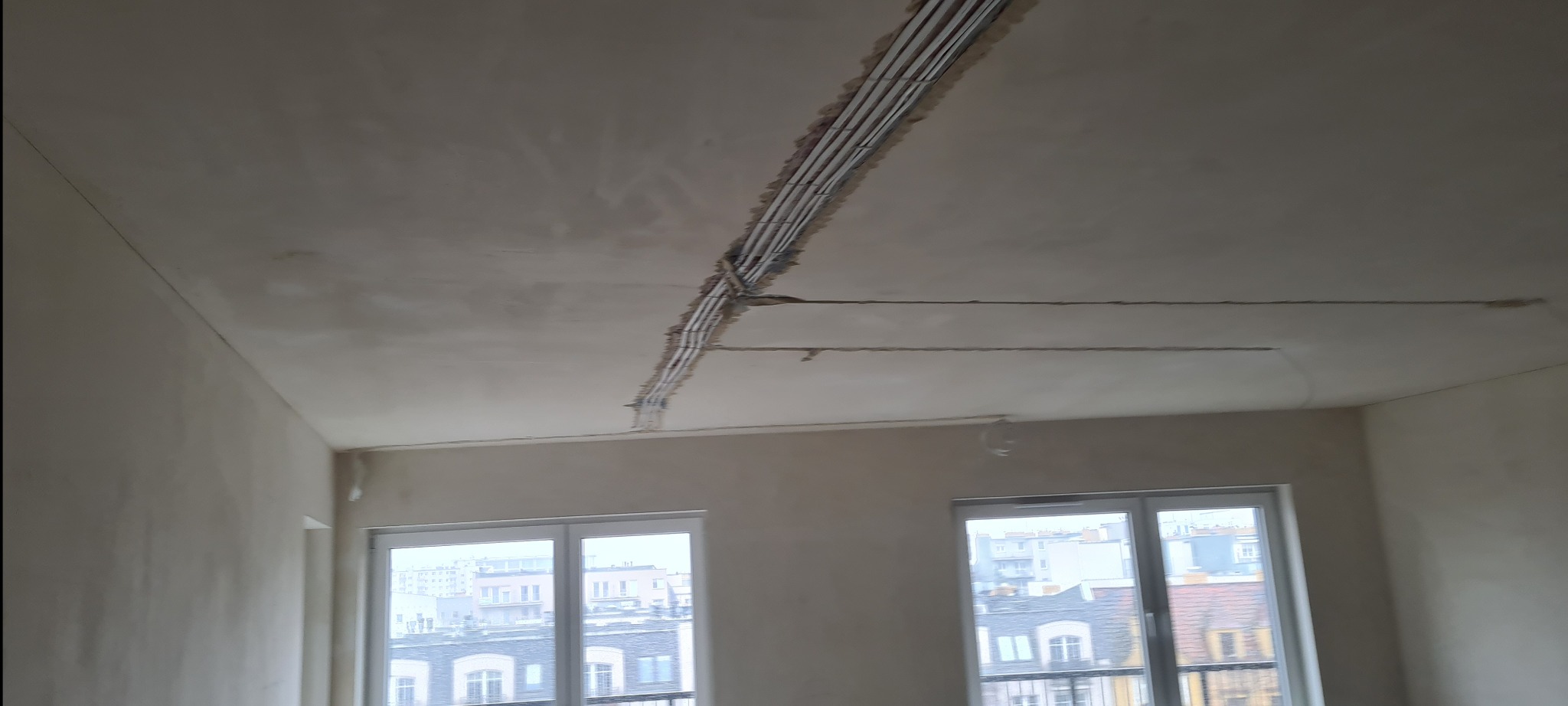 Wnętrze w stanie deweloperskim z widoczną instalacją elektryczną w bruzdach na suficie, przed położeniem tynku. Dwa okna z widokiem na budynki miejskie.