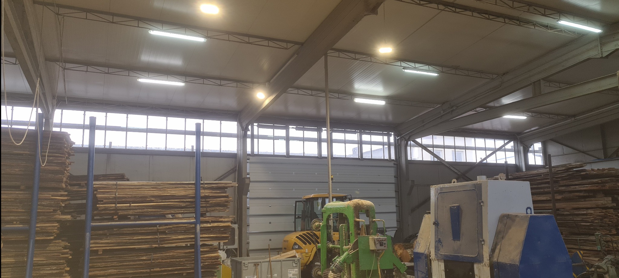 Oświetlenie LED w hali produkcyjnej z maszynami do obróbki drewna i składowanym drewnem. Widoczne lampy jarzeniowe i punktowe LED na suficie.