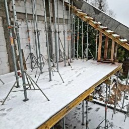 KECHARIT Tomasz Baranowski - Szare stemple budowlane podtrzymują drewniany szalunek stropu, pokryty śniegiem. Widoczne ślady stóp. Ściany z białych bloczków.