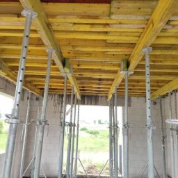KECHARIT Tomasz Baranowski - Żółte szalunki stropowe podparte metalowymi stemplami w surowym wnętrzu. Widok z dołu na konstrukcję nośną stropu w trakcie prac budowlanych.
