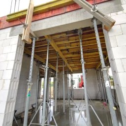 KECHARIT Tomasz Baranowski - Wnętrze budynku w trakcie budowy z białych bloczków, wsparte metalowymi stemplami i drewnianymi belkami, widoczny czerwony żuraw za oknem.