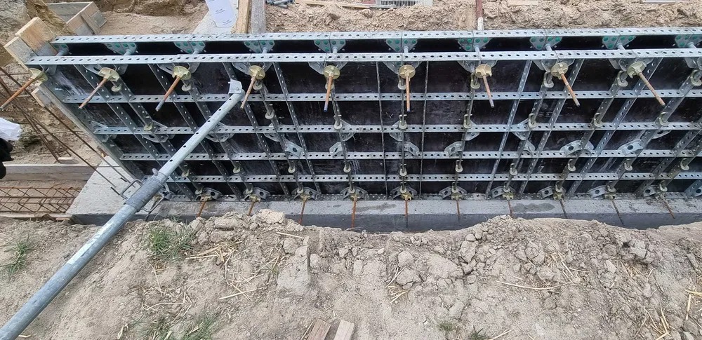Szczegółowe ujęcie szalunku fundamentowego z widocznymi elementami mocującymi i podporą teleskopową, na tle wykopu zbrojeniowego.