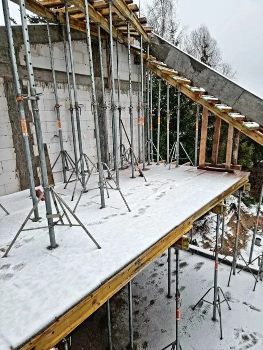 Szare stemple budowlane podtrzymują drewniany szalunek stropu, pokryty śniegiem. Widoczne ślady stóp. Ściany z białych bloczków.