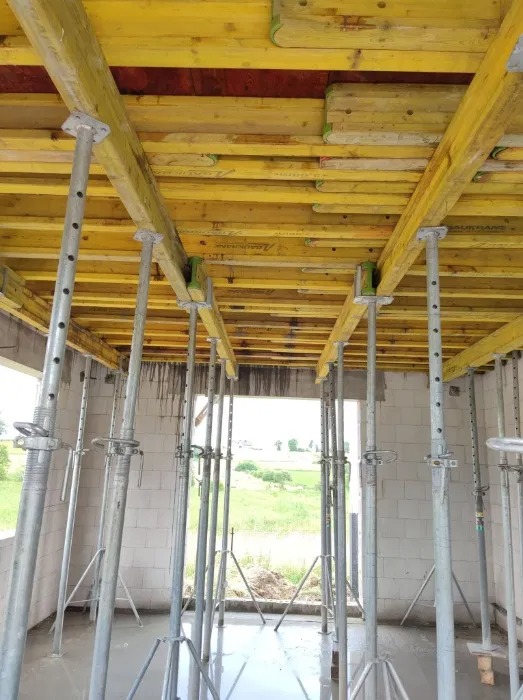 Żółte szalunki stropowe podparte metalowymi stemplami w surowym wnętrzu. Widok z dołu na konstrukcję nośną stropu w trakcie prac budowlanych.