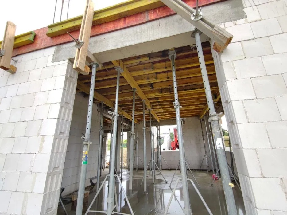 Wnętrze budynku w trakcie budowy z białych bloczków, wsparte metalowymi stemplami i drewnianymi belkami, widoczny czerwony żuraw za oknem.