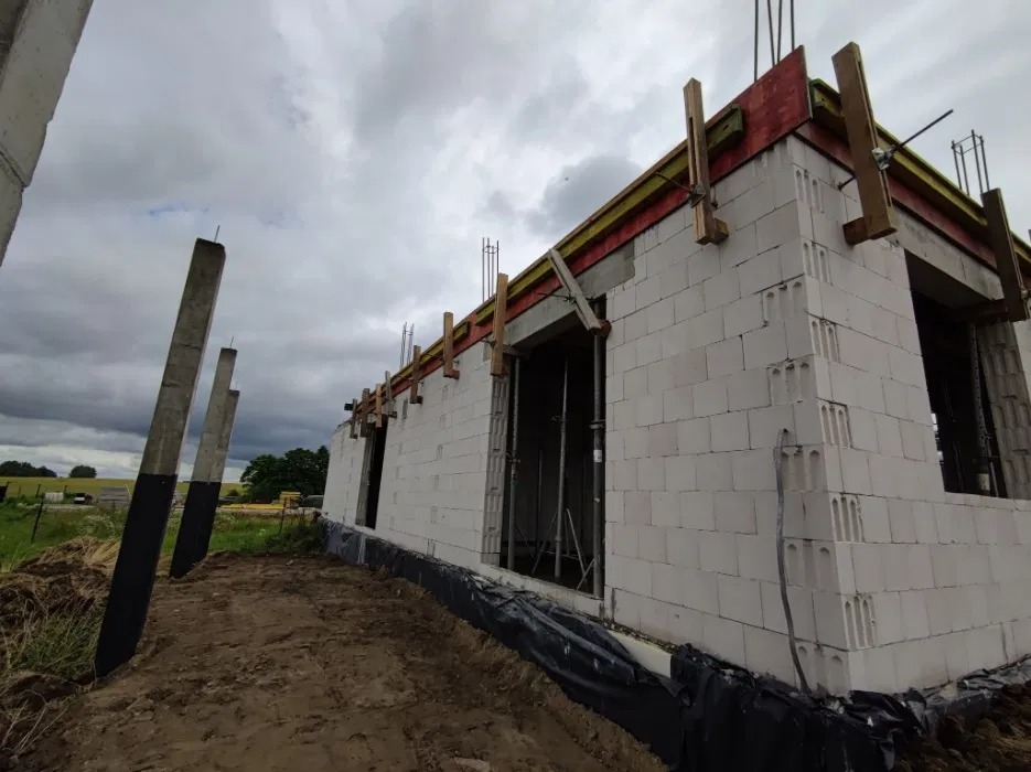 Budowa domu z betonu komórkowego w trakcie realizacji, widoczne ściany, szalunki i podpory. Prace murarskie w toku, teren budowy, pochmurne niebo.