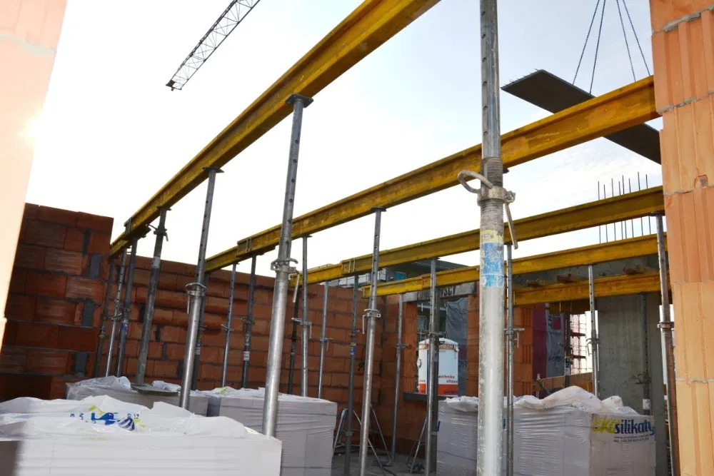 Wnętrze budowy z żółtymi dźwigarami i stalowymi podporami. Na pierwszym planie palety z cegłami silikatowymi. W tle widoczny żuraw budowlany.