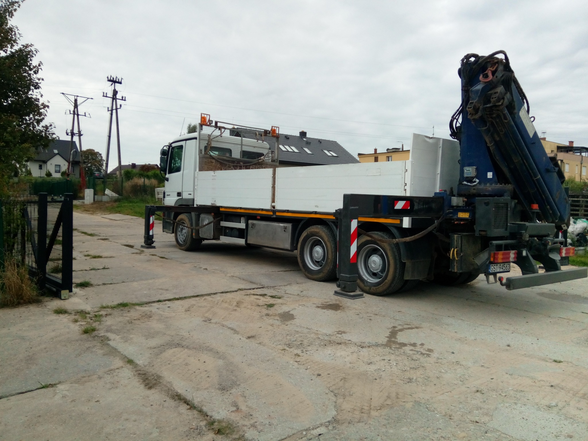 Ciężarówka z dźwigiem hydraulicznym zaparkowana na betonowym placu, widoczna brama wjazdowa i słupy energetyczne w tle. Dźwig złożony, gotowy do transportu.