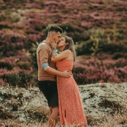 Czułe Światło - Zakochana para całuje się na tle wrzosowiska. Mężczyzna z tatuażem na ramieniu obejmuje kobietę w kwiecistej sukience. Romantyczna sesja w plenerze.