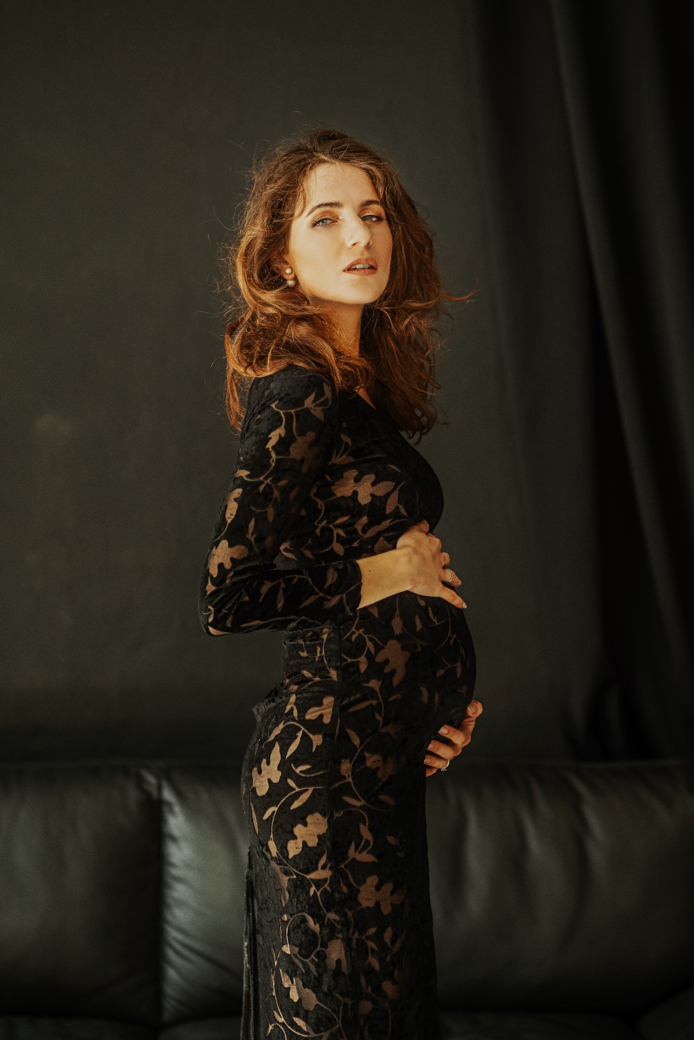 Kobieta w czarnej, koronkowej sukience ciążowej, pozująca na tle ciemnego studia z skórzaną kanapą; delikatny makijaż, subtelna biżuteria, profesjonalne oświetlenie.
