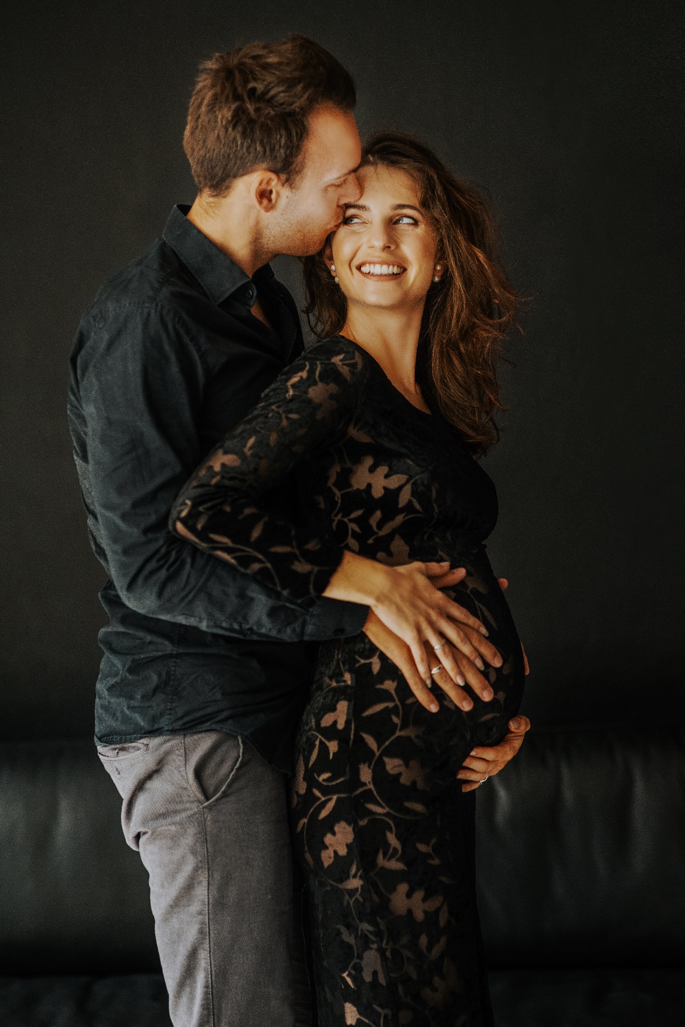 Sesja ciążowa w studio: mężczyzna całuje w głowę uśmiechniętą kobietę w czarnej, kwiecistej sukni, trzymającą dłonie na brzuchu na tle ciemnego tła.