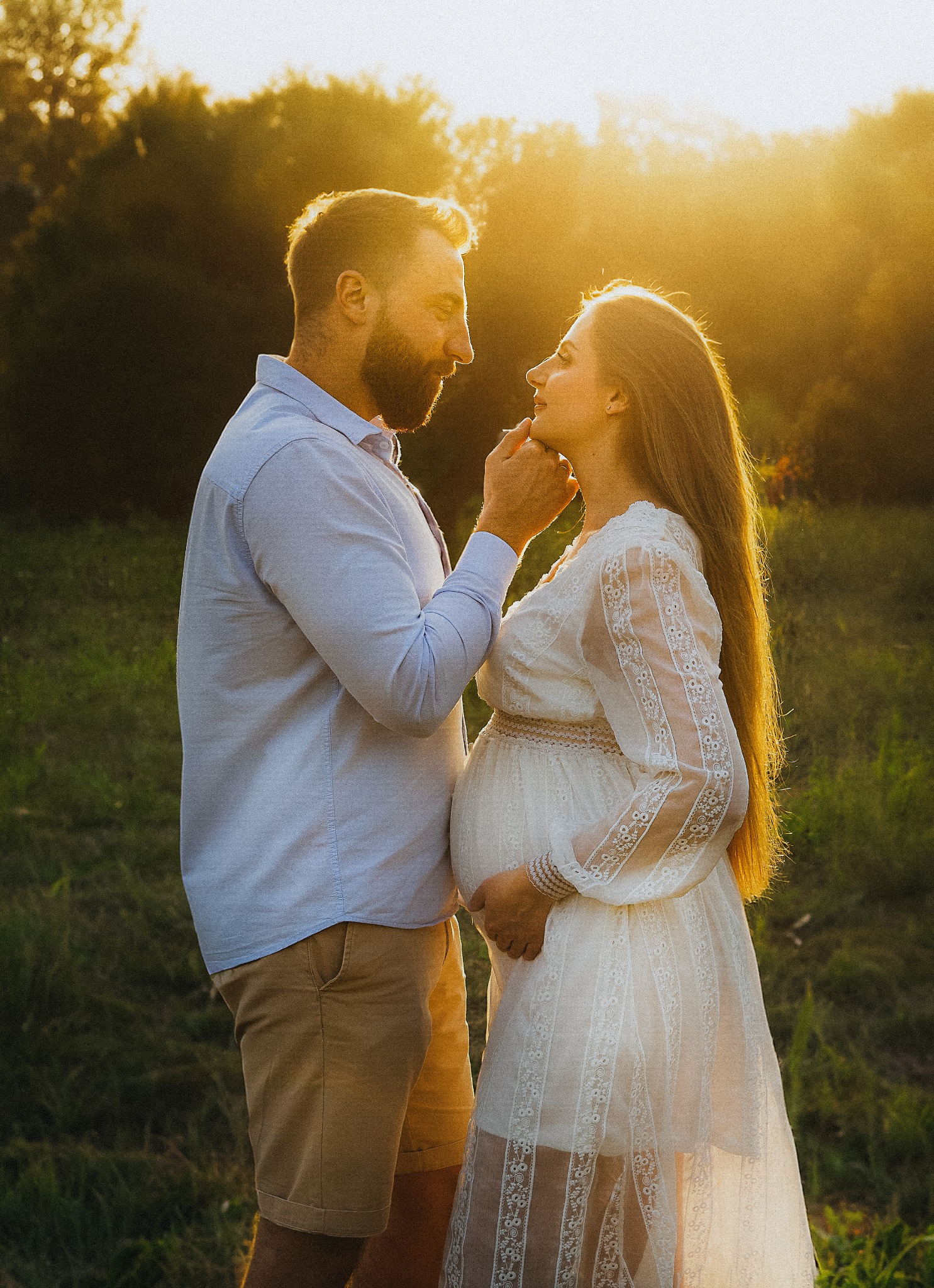 Para w plenerze, kobieta w zwiewnej sukience ciążowej, mężczyzna dotyka jej twarzy, ciepłe światło zachodzącego słońca, naturalne tło.