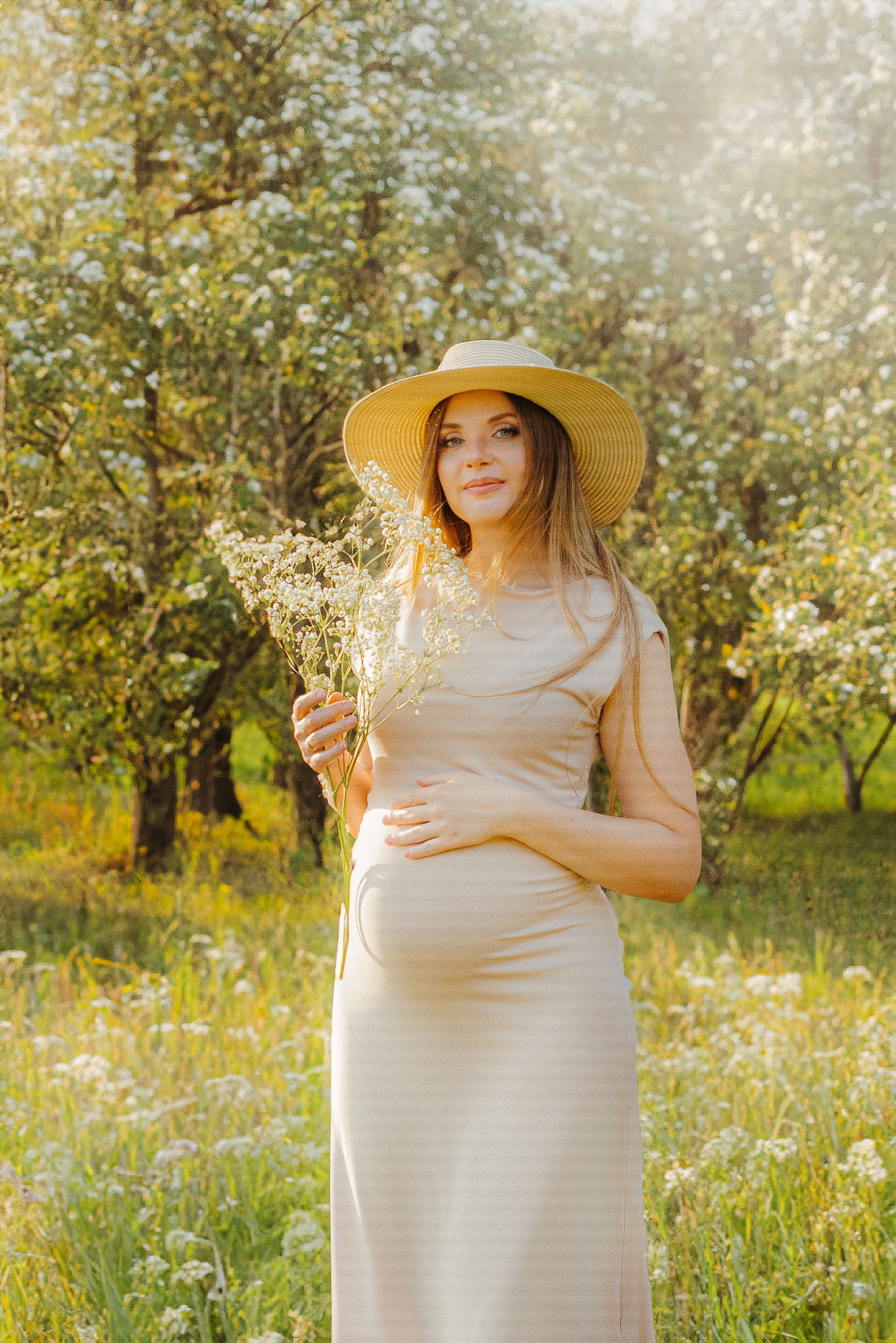 Kobieta w ciąży, w beżowej sukience i kapeluszu, pozuje w plenerze z białymi kwiatami na tle kwitnących drzew, w słonecznym świetle.