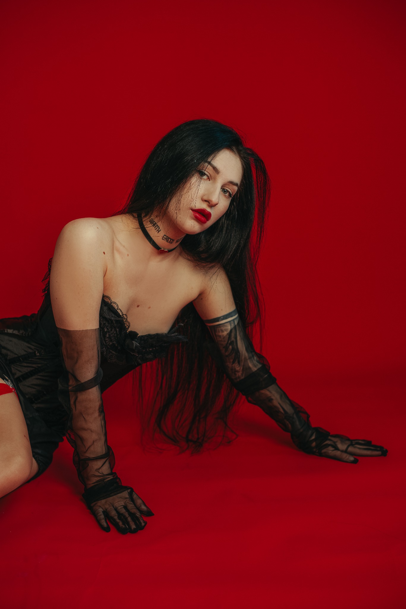 Kobieta w czarnej sukience i rękawiczkach na czerwonym tle. Uwagę przykuwa choker z napisem oraz intensywna czerwień ust. Ujęcie z góry podkreśla pozę modelki.