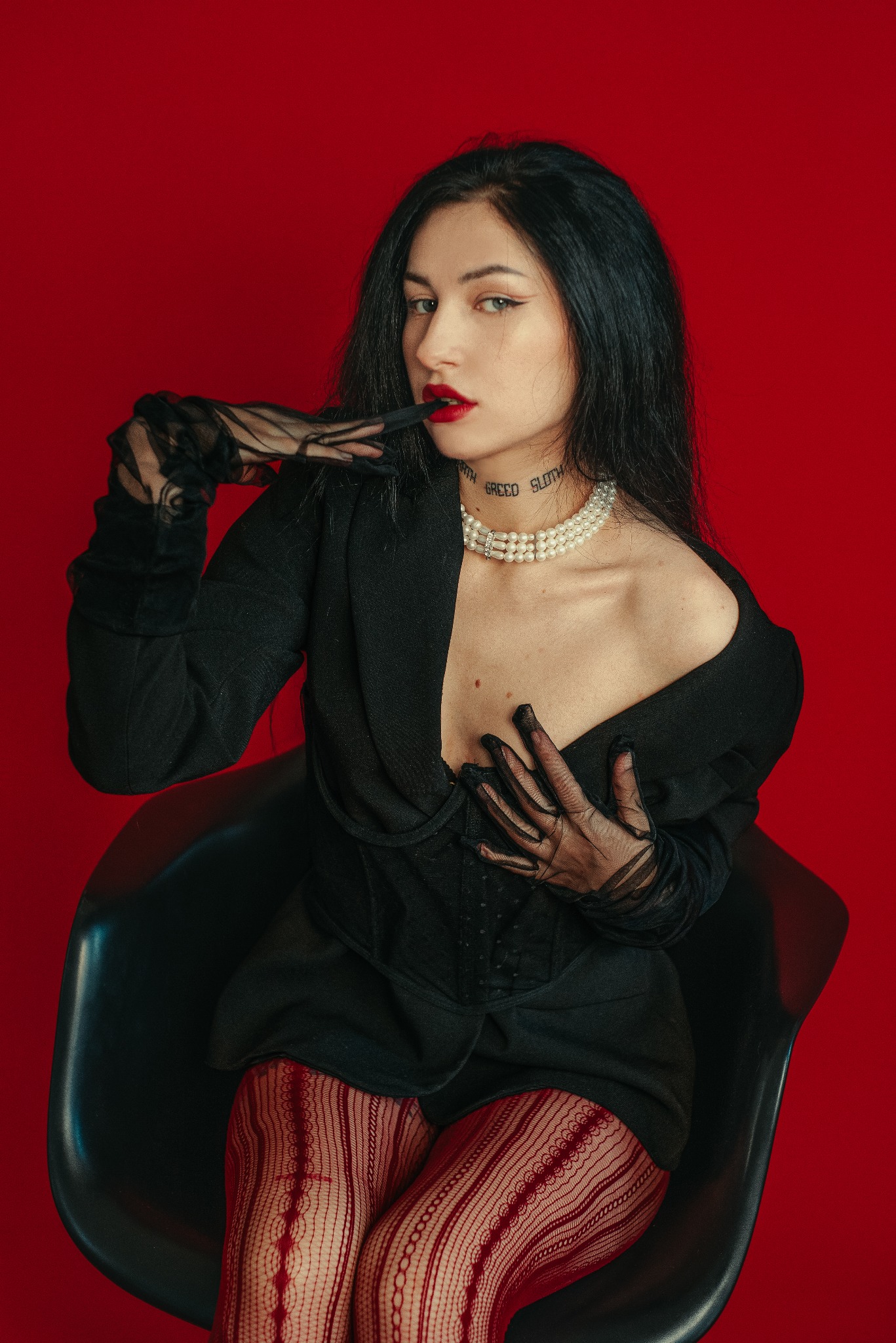 Kobieta w czarnym żakiecie, perłach i czerwonych rajstopach pozuje na krześle na czerwonym tle. Usta pomalowane czerwoną szminką, na szyi widoczny tatuaż.