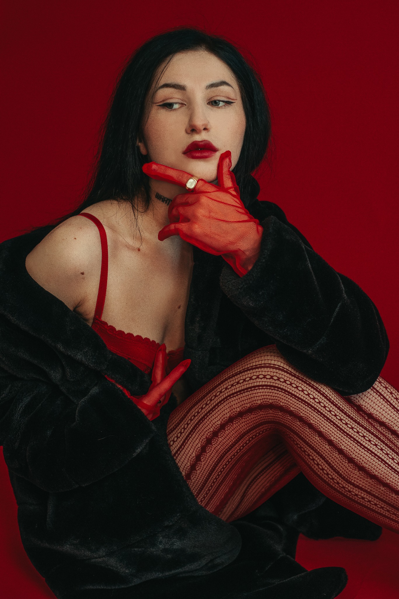 Kobieca sesja w studio: Brunetka w czerwonej bieliźnie, kabaretkach i futrze na tle czerwonego tła. Uwagę przykuwa pierścionek na palcu.