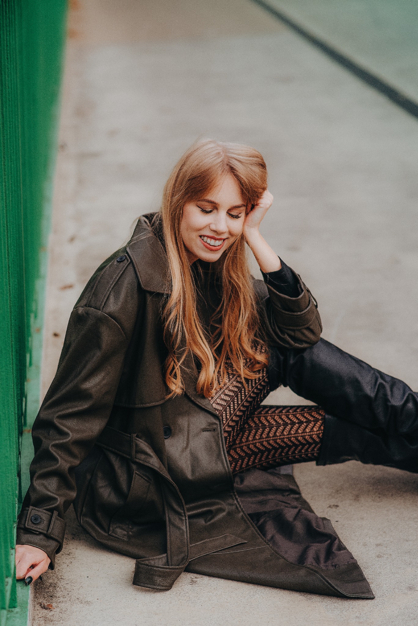 Uśmiechnięta blondynka w skórzanym płaszczu siedzi na szarym tle obok zielonej barierki, opierając głowę na dłoni. Ubrana w rajstopy we wzory i czarną bluzkę.
