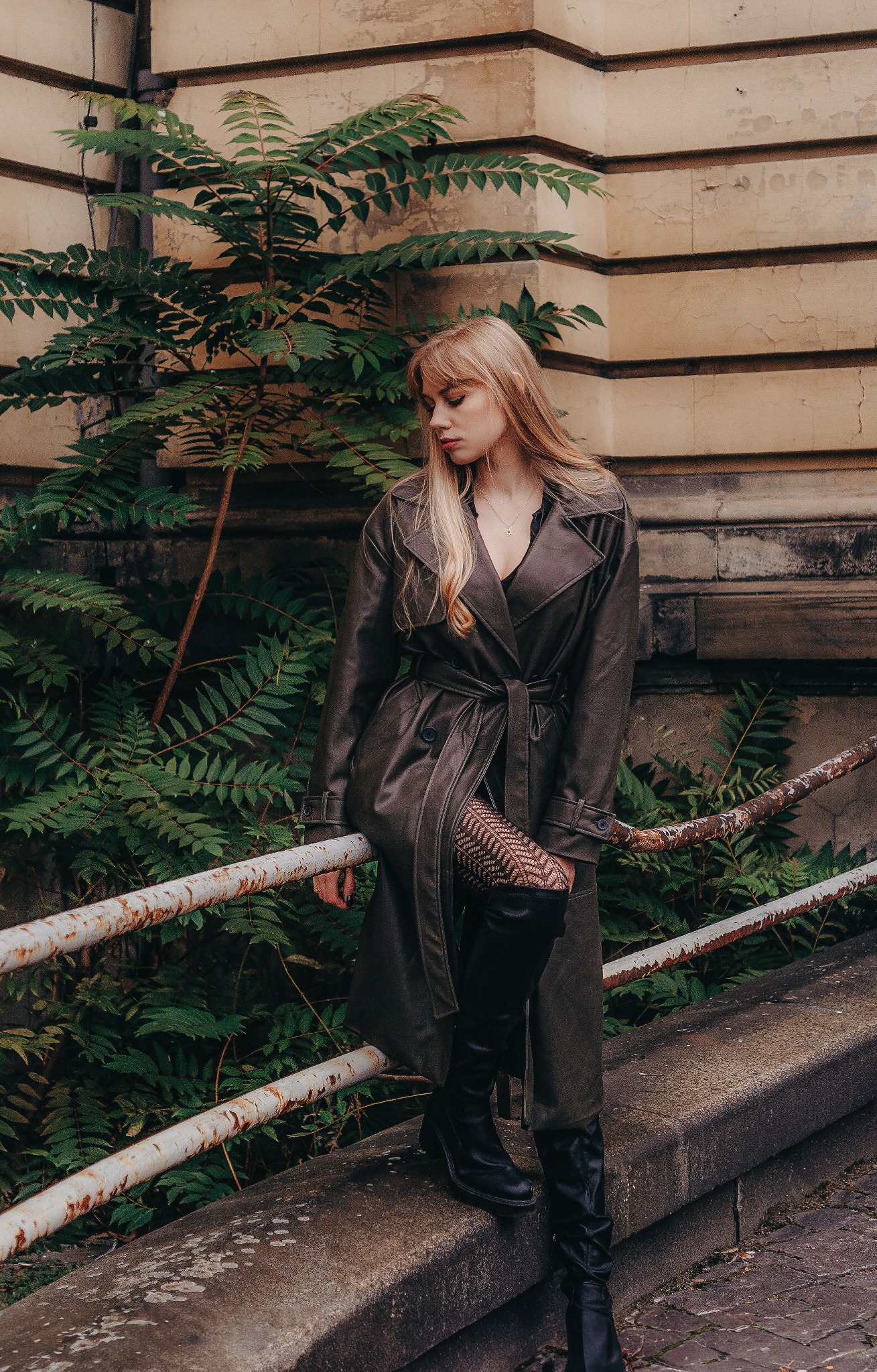 Plenerowa sesja: kobieta w płaszczu siedzi na barierce, za nią zielona roślina i budynek. Uwagę przykuwają czarne, wysokie buty i rajstopy w delikatny wzór.