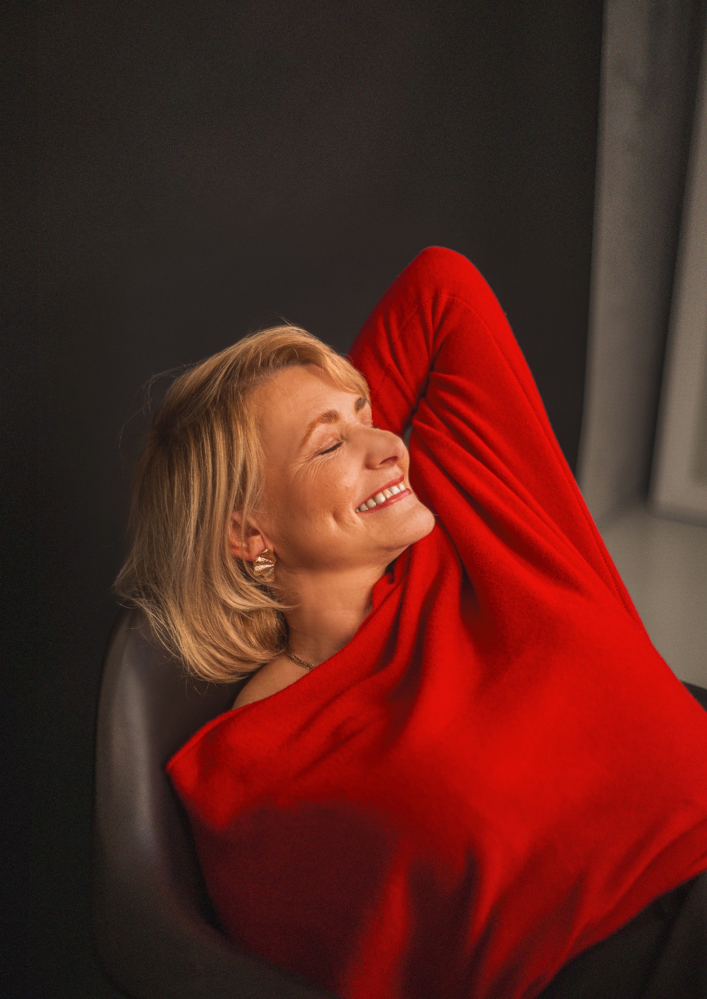 Portret uśmiechniętej kobiety w czerwonym swetrze, z zamkniętymi oczami, w studio na ciemnym tle, z widocznym oknem po prawej stronie.