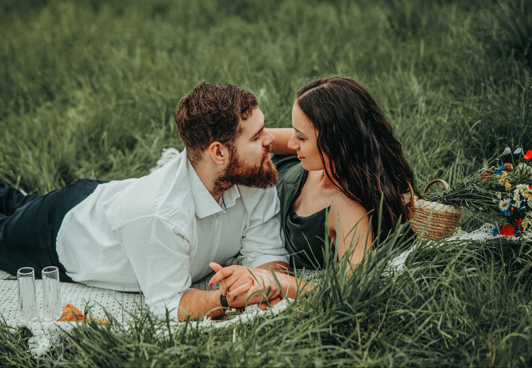 Zakochana para leży na kocu w trawie, trzymając się za ręce. Koszyk z kwiatami i szklanki dopełniają romantyczną scenerię sesji plenerowej.