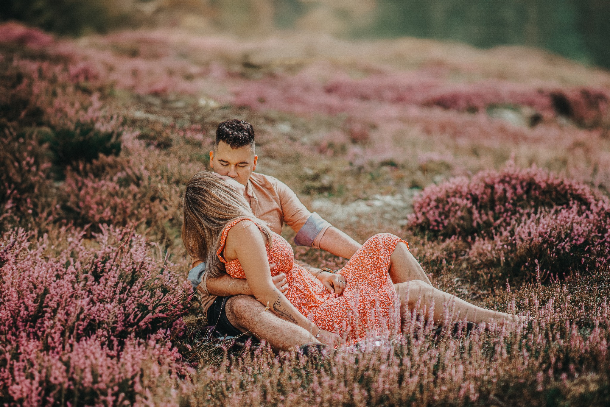 Zakochana para w objęciach siedzi na wrzosowisku podczas sesji narzeczeńskiej, mężczyzna całuje kobietę w głowę. Romantyczna atmosfera w plenerze.