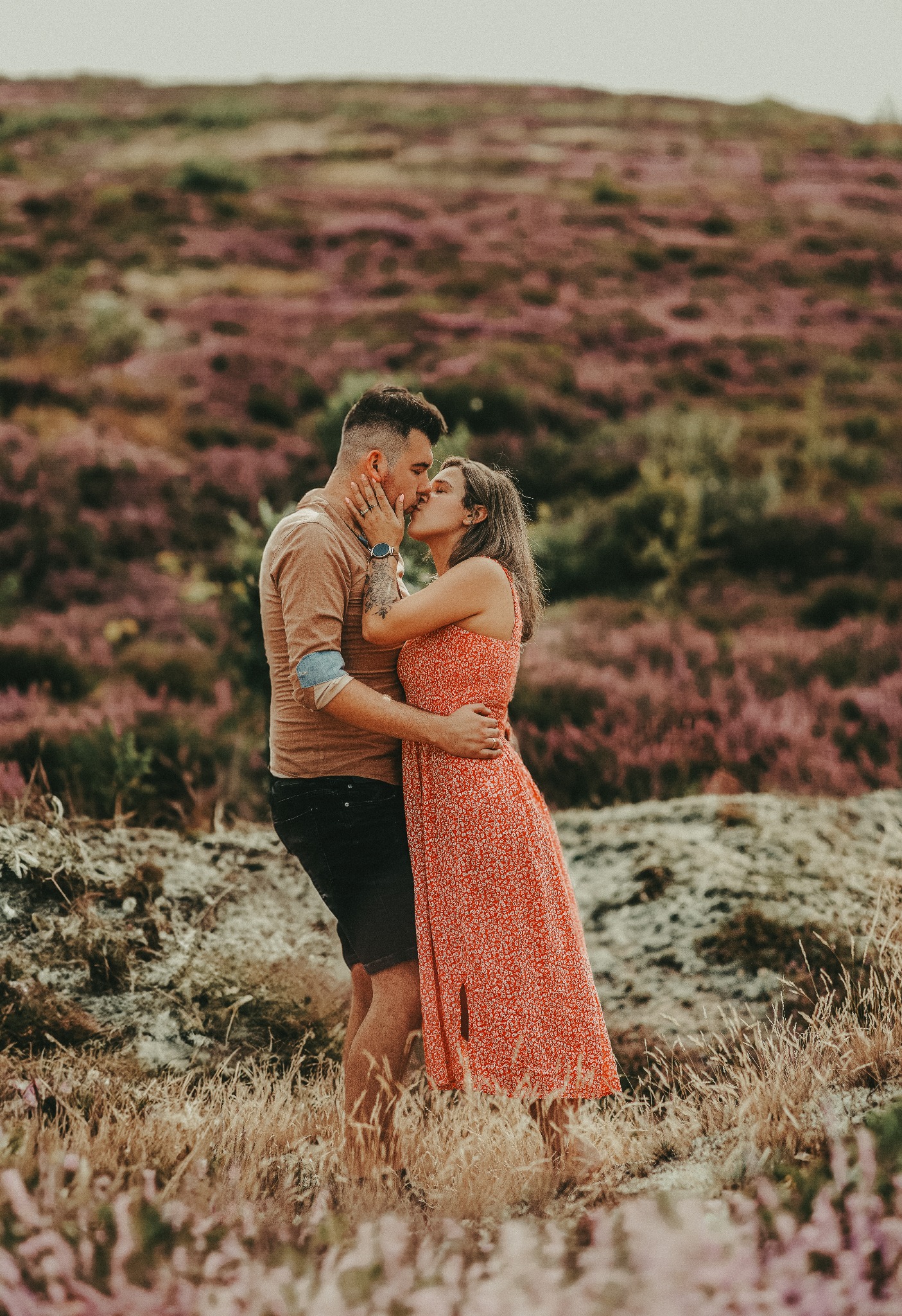 Zakochana para całuje się na tle wrzosowiska. Mężczyzna z tatuażem na ramieniu obejmuje kobietę w kwiecistej sukience. Romantyczna sesja w plenerze.