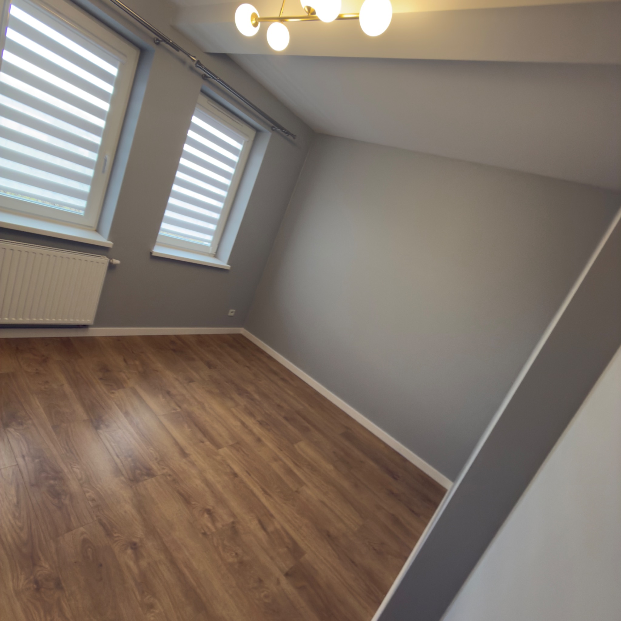 Jasne, szare wnętrze po remoncie: drewniana podłoga, grzejnik pod oknem z roletami, nowoczesna lampa sufitowa. Ściany pomalowane na szaro.