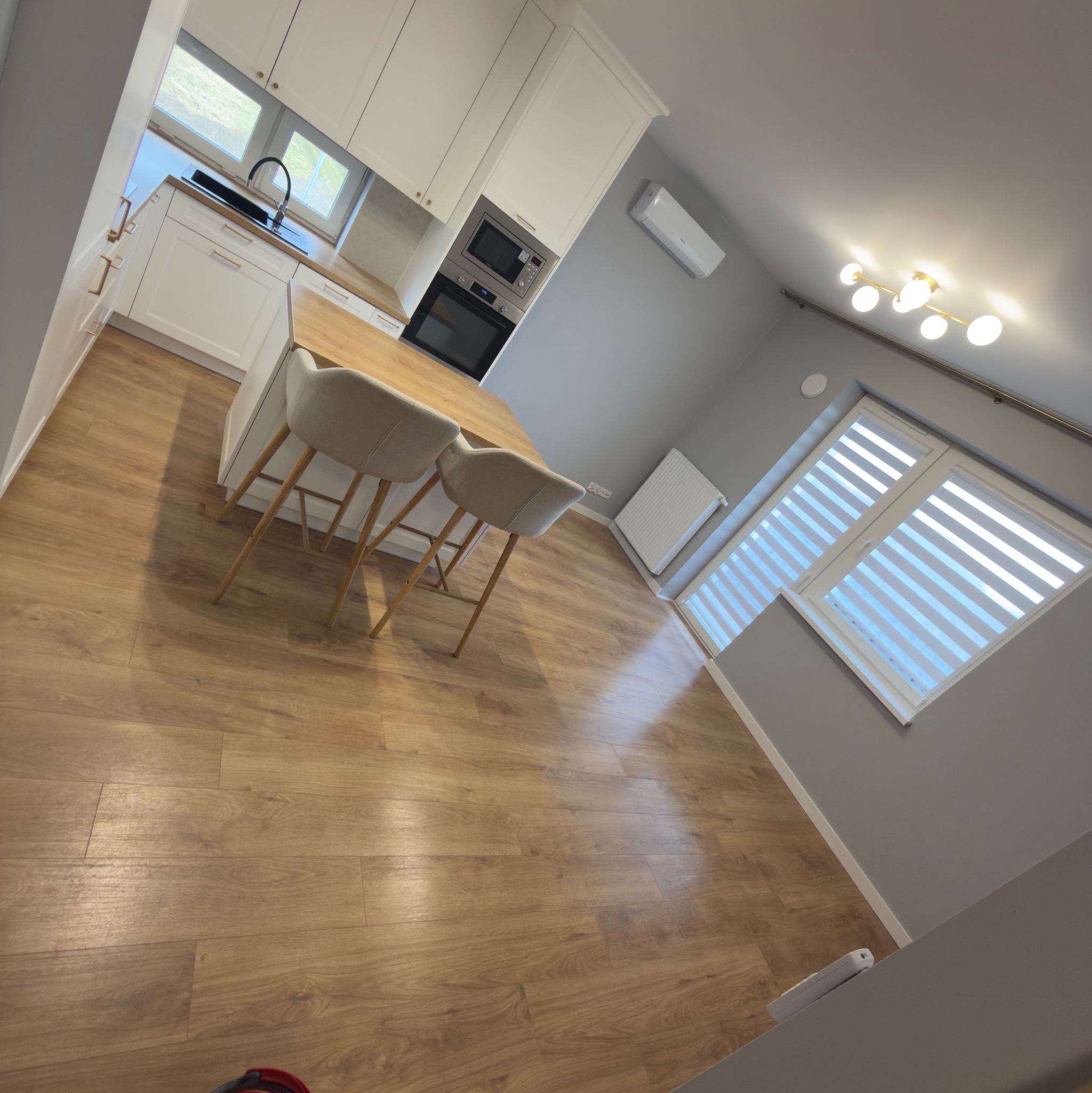 Jasne, nowocześnie wykończone wnętrze z drewnianą podłogą, białymi meblami kuchennymi i wyspą z hokerami. Widoczne okna z żaluzjami i designerska lampa sufitowa.