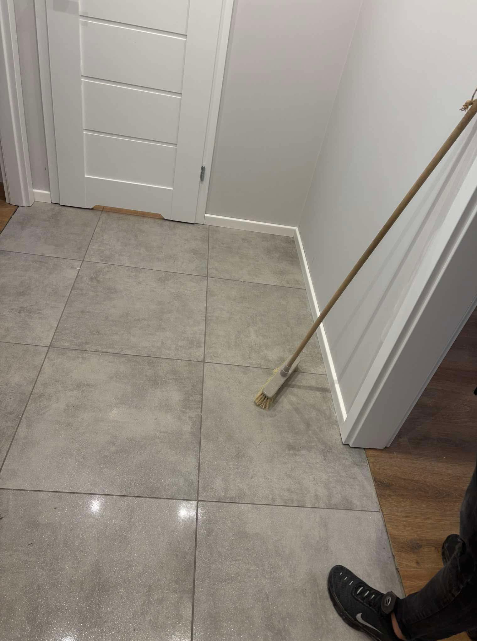 Szare płytki podłogowe w korytarzu, częściowo widoczna biała ściana i drzwi. Na podłodze stoi miotła, a w rogu widać fragment buta.