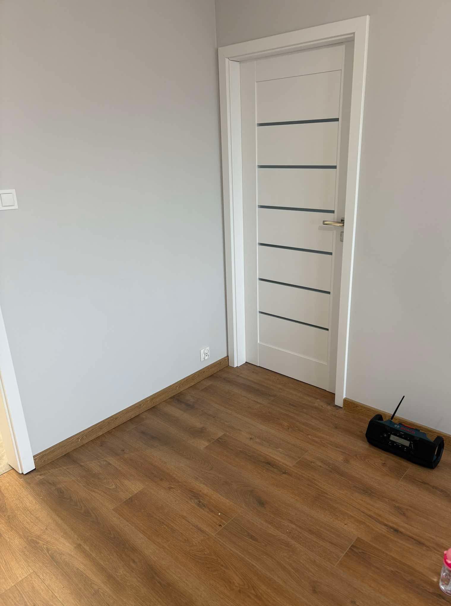 Świeżo wykończone wnętrze: szare ściany, drewnopodobna podłoga i białe drzwi z poziomymi, szarymi paskami. Radio budowlane leży na podłodze.