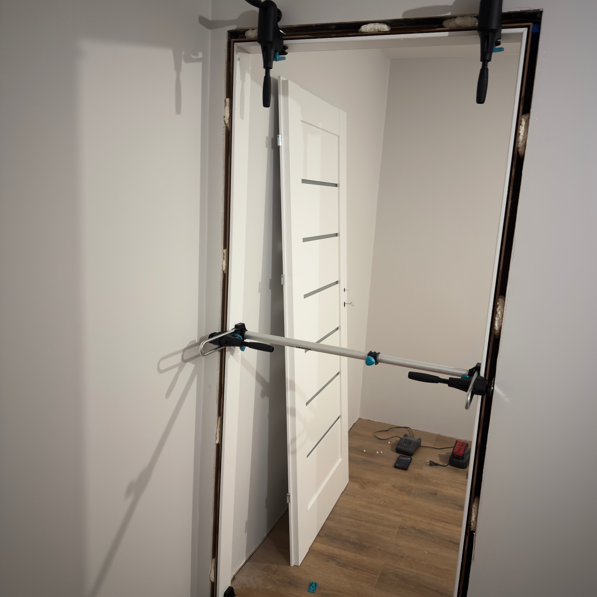 Montaż białych drzwi wewnętrznych z poziomymi frezami. Użycie rozpórki montażowej. Widoczne pianka montażowa i narzędzia na podłodze. Ujęcie z bliska, w trakcie pracy.