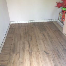 Re Monty - Ukończona podłoga z paneli imitujących drewno, z białymi płytkami przypodłogowymi. W tle zabawki dziecięce,  widok z perspektywy podłogi.