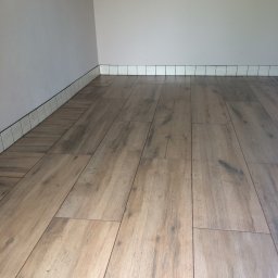 Re Monty - Ułożona podłoga z paneli imitujących drewno, przy ścianie białe płytki cokołowe z ciemną fugą. Widok na fragment wykończonego wnętrza.