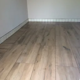 Ułożona podłoga z paneli imitujących drewno, przy ścianie białe płytki cokołowe z ciemną fugą. Widok na fragment wykończonego wnętrza.