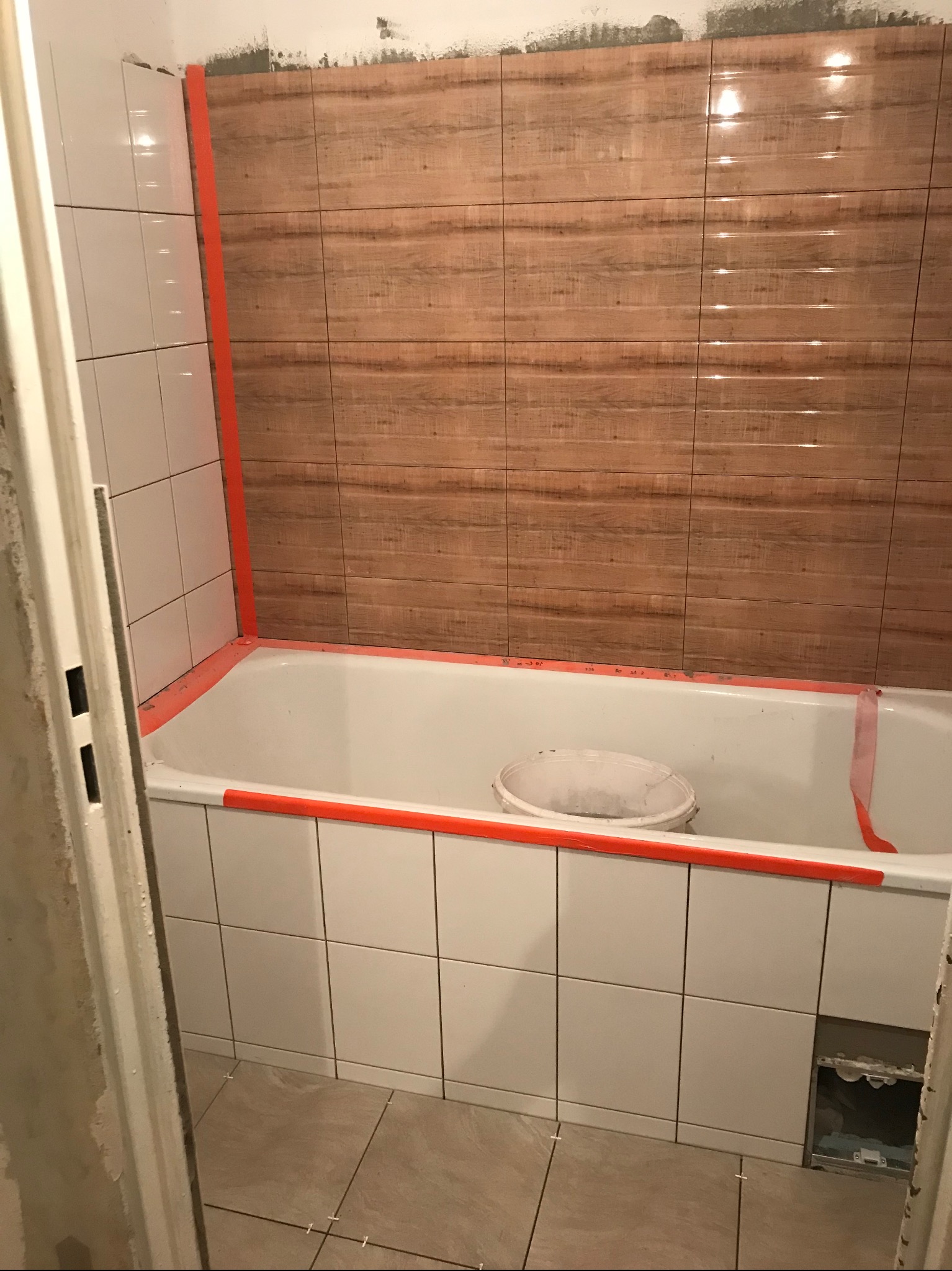 Łazienka w trakcie prac: białe i drewnopodobne płytki, wanna obłożona kafelkami, widoczna taśma zabezpieczająca i wiadro. Częściowo wykończone wnętrze.