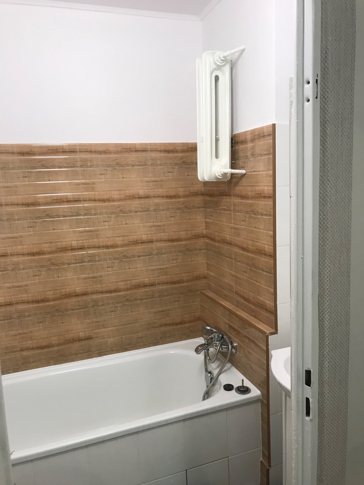 Biała wanna z baterią i brązowe płytki imitujące drewno na ścianie łazienki. Widoczny grzejnik i fragment umywalki. Minimalistyczny styl.