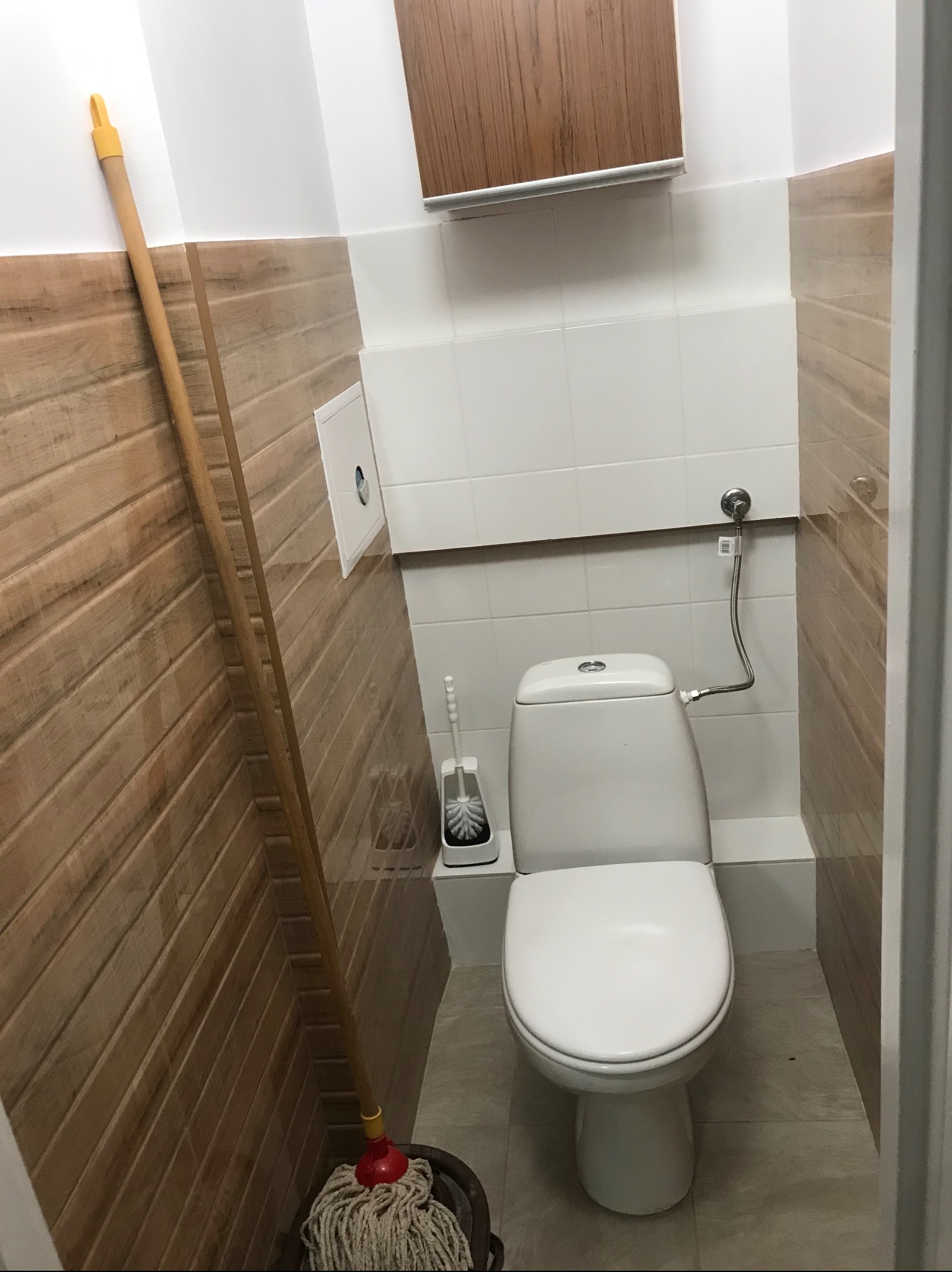 Biała toaleta w łazience z drewnopodobnymi panelami ściennymi i mopem. Widoczna instalacja wodna i szafka wisząca. Ukończone wykończenie wnętrza łazienki.