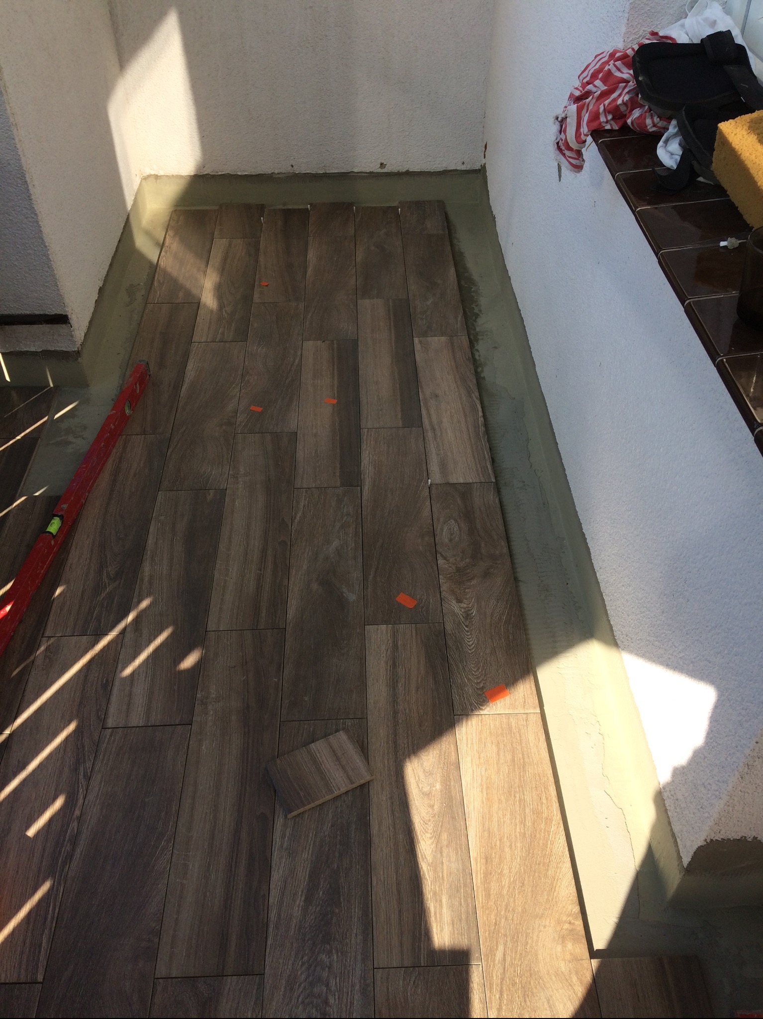 Układanie drewnopodobnych płytek na balkonie. Widoczny poziomica, krzyżyki dystansowe i resztki zaprawy. W tle ściana z fragmentem półki.