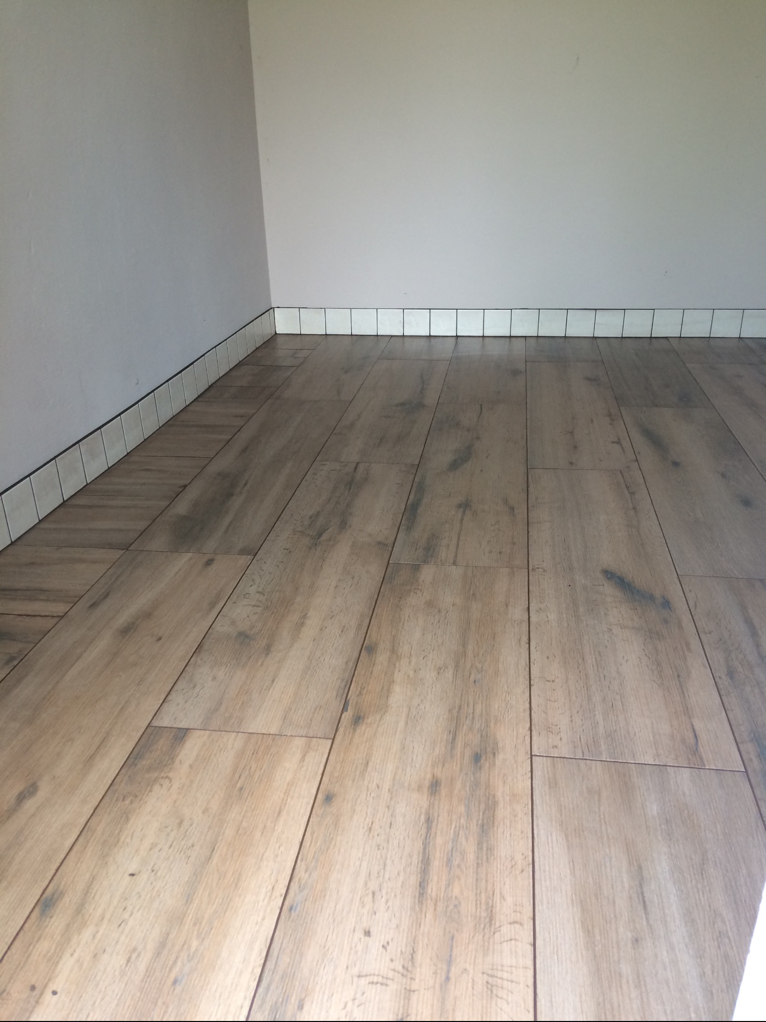 Ułożona podłoga z paneli imitujących drewno, przy ścianie białe płytki cokołowe z ciemną fugą. Widok na fragment wykończonego wnętrza.