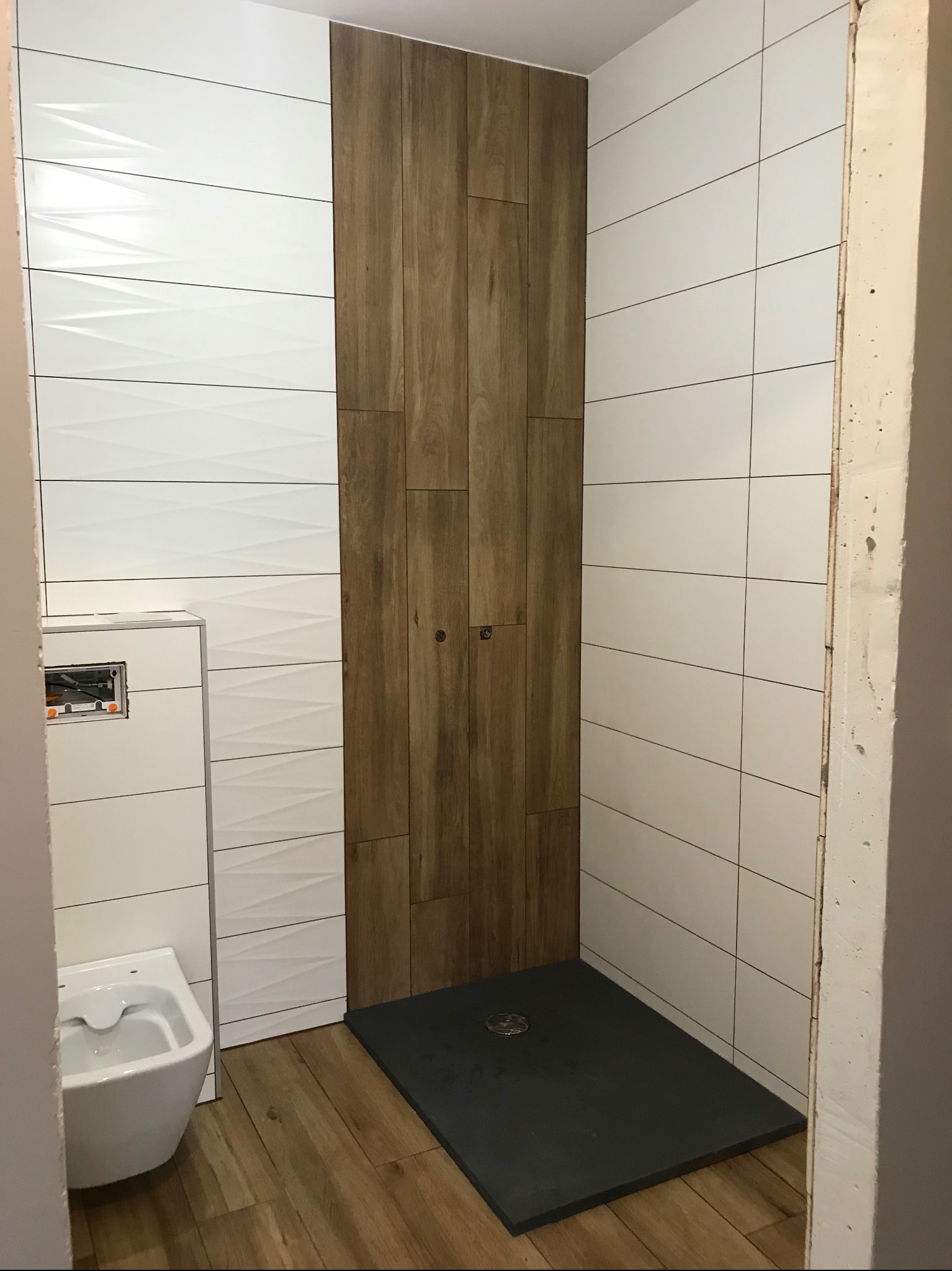 Nowoczesna kabina prysznicowa z drewnopodobnymi i białymi płytkami. Widoczny odpływ liniowy, bidet i fragment surowej ściany.