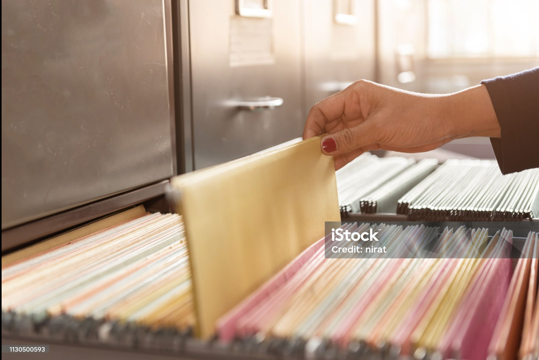 Otwarta szafa kartotekowa z kolorowymi teczkami, dłoń wkłada beżową teczkę. Organizacja dokumentów w biurze, archiwizacja danych, zarządzanie informacjami.