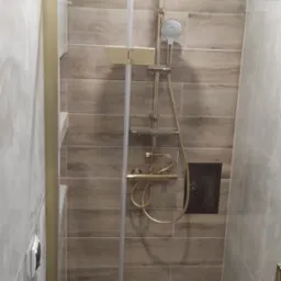 Wykończona kabina prysznicowa z deszczownicą w odcieniach beżu i złota. Widoczne płytki imitujące drewno, szklane drzwi z złotymi okuciami i punktowe oświetlenie sufitowe.