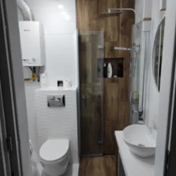 Nowoczesna łazienka z drewnianymi akcentami: kabina prysznicowa z deszczownicą, umywalka nablatowa i podwieszana toaleta. Białe płytki z fakturą i piec Junkers.