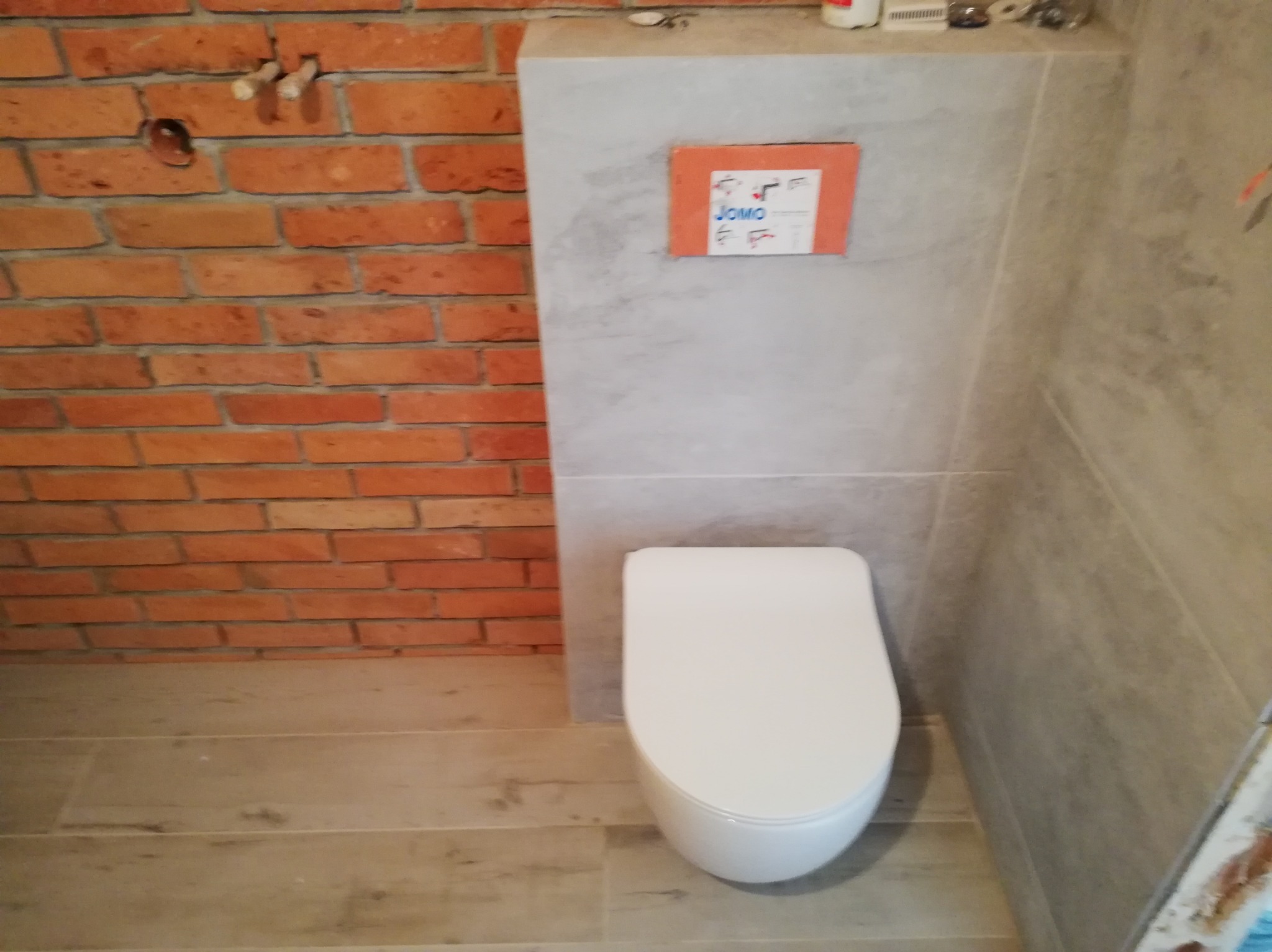 Nowoczesna toaleta podwieszana na tle ściany z cegły i płytek imitujących beton. Widoczne elementy instalacji. Minimalistyczny design z akcentem industrialnym.