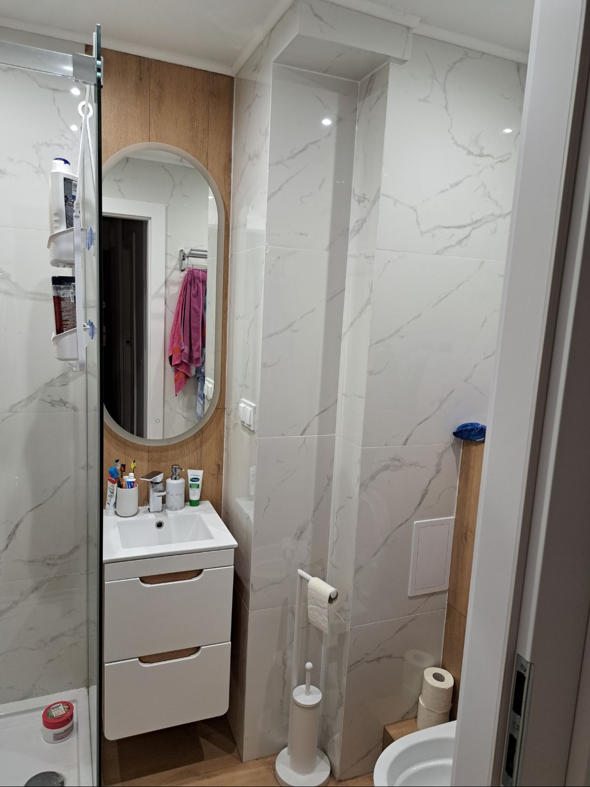 Wykończona łazienka z białą armaturą i płytkami imitującymi marmur. Umywalka z szafką, owalne lustro i toaleta. Widoczne detale wykończenia.