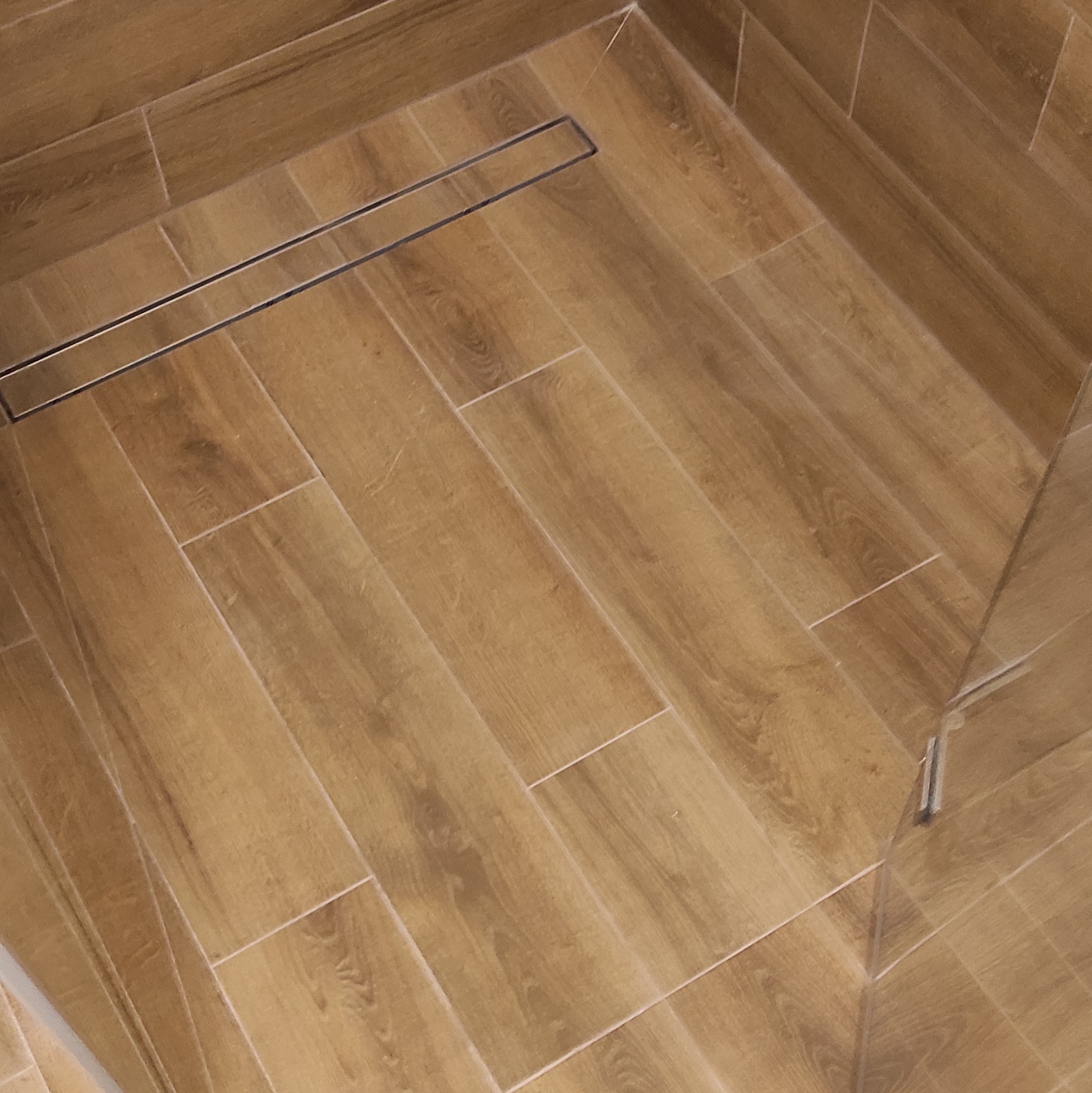 Ukończona podłoga w łazience z drewnopodobnych płytek ułożonych w jodełkę, z odpływem liniowym. Widoczny fragment szklanej kabiny prysznicowej.