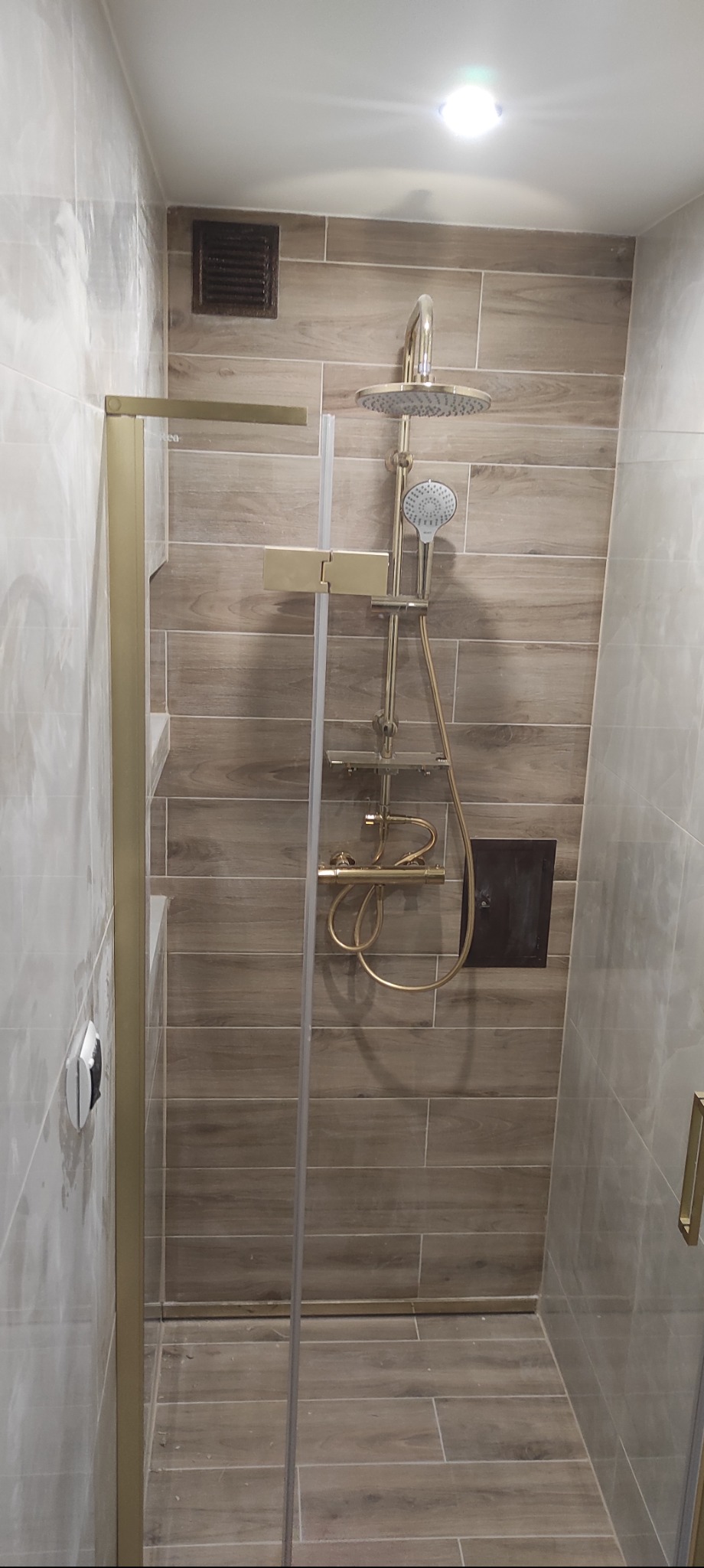 Wykończona kabina prysznicowa z deszczownicą w odcieniach beżu i złota. Widoczne płytki imitujące drewno, szklane drzwi z złotymi okuciami i punktowe oświetlenie sufitowe.