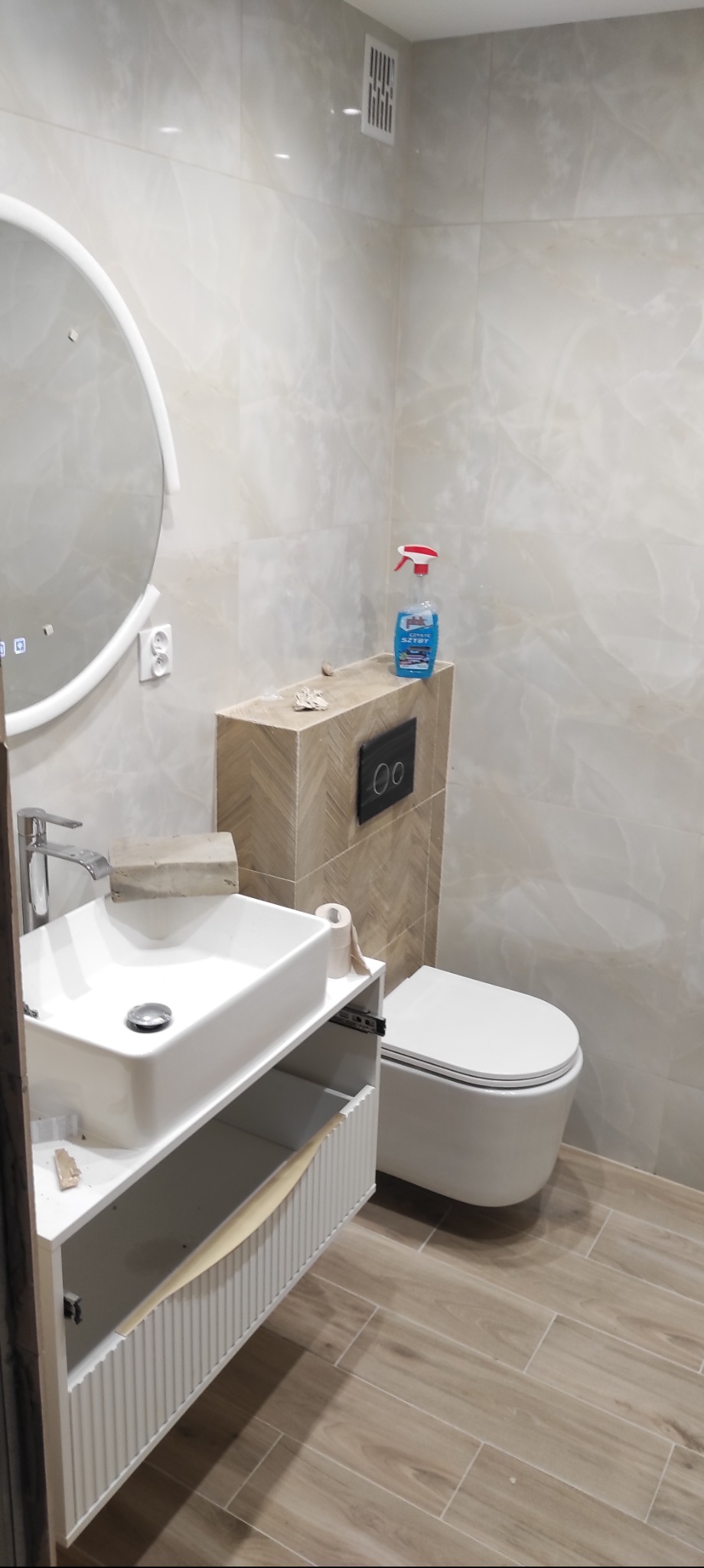 Nowoczesna łazienka z podwieszaną toaletą, umywalką na szafce z otwartą szufladą i okrągłym lustrem z podświetleniem LED. Płytki imitujące marmur na ścianach i podłodze.